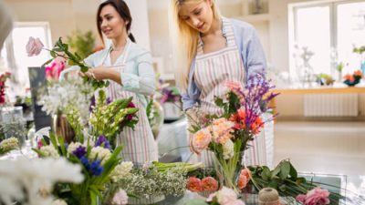 Two women wearing striped aprons are arranging colorful flower bouquets in a bright, spacious room filled with natural light and various floral displays.