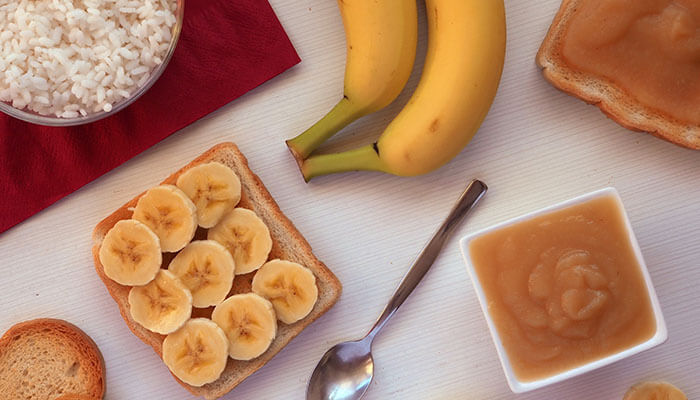 Slices of banana on toast, a bowl of applesauce, plain rice, and bananas displayed on a table, representing foods from the brat or bland diet.