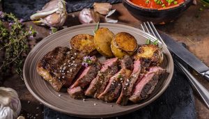 A plate of grilled steak slices, part of carnivore diet recipes, served with roasted potatoes, garnished with herbs and sea salt, and accompanied by garlic and fresh vegetables in the background.