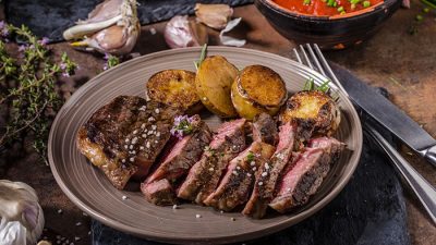 A plate of grilled steak slices, part of carnivore diet recipes, served with roasted potatoes, garnished with herbs and sea salt, and accompanied by garlic and fresh vegetables in the background.