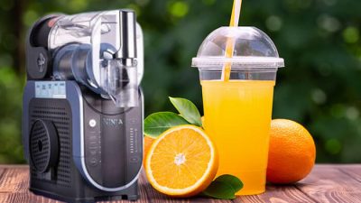 Ninja Slushie machine with a fresh orange slushie in a cup, surrounded by oranges on a wooden table.