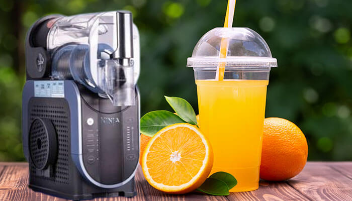 Ninja Slushie machine with a fresh orange slushie in a cup, surrounded by oranges on a wooden table.