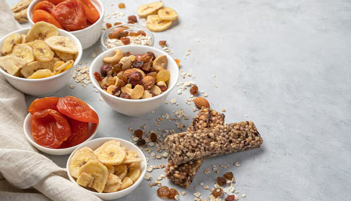 Assorted high protein snacks including dried fruits, nuts, and protein bars displayed in bowls on a light background.