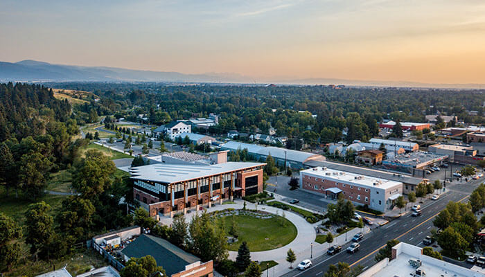 Aerial view of Missoula, Montana, at sunset featuring the University of Montana campus buildings, tree-lined streets, and distant mountain ranges under a warm golden sky.
