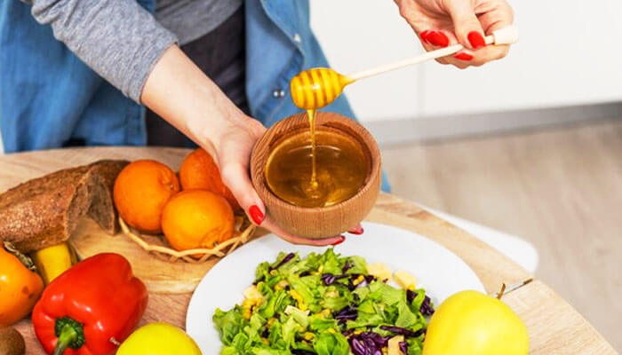 Woman pouring honey over a fresh salad surrounded by fruits and vegetables, illustrating the dietary question — is honey vegan or not.