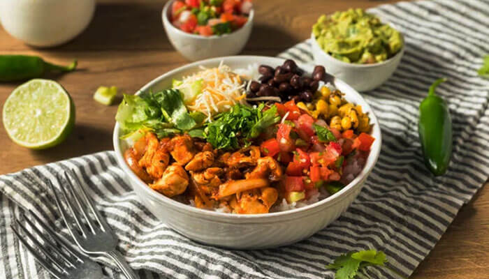 Colorful burrito bowl filled with grilled chicken, rice, beans, corn, salsa, cheese, and guacamole, served on a striped cloth with jalapeños and lime — a healthy Mexican meal.