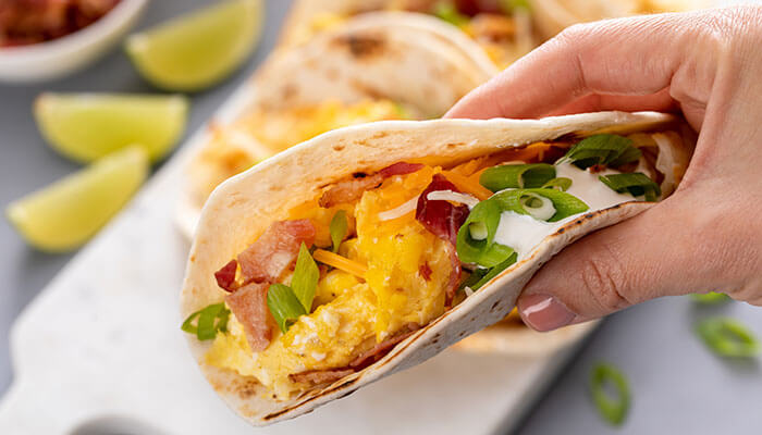 Hand holding a breakfast taco filled with scrambled eggs, bacon, cheese, sour cream, and green onions, with lime wedges in the background — a quick morning meal.