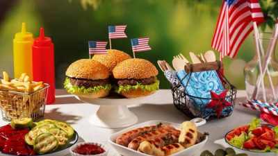 Table filled with classic American cuisine including burgers, hot dogs, fries, and sauces decorated with small U.S. flags for a festive meal.