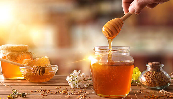 Golden honey dripping from a dipper into a glass jar on a wooden table — a visual symbol for the ethical debate around is honey vegan.