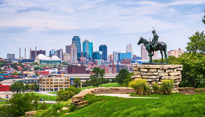 Scenic view of Kansas City, Missouri skyline with the iconic Scout statue in the foreground, overlooking modern skyscrapers, green parks, and a vibrant downtown area under a partly cloudy sky.