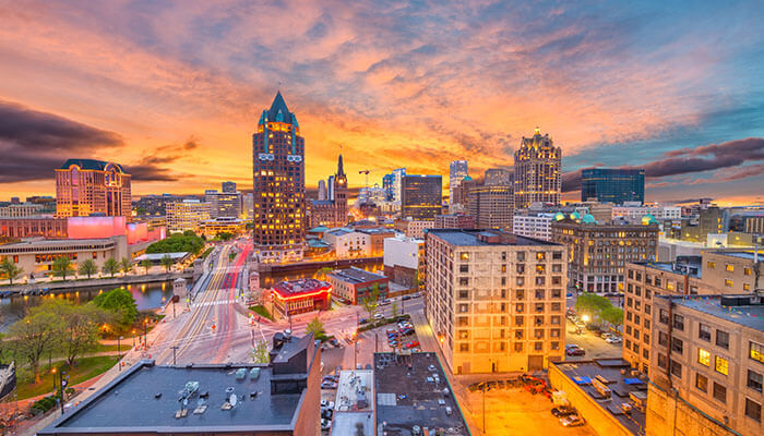 Vibrant sunset view of downtown Milwaukee, Wisconsin, showcasing illuminated skyscrapers, the Milwaukee River, and colorful city lights reflecting against a dramatic evening sky.