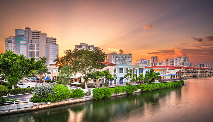 Scenic view of Naples, Florida at sunset, featuring waterfront homes, lush greenery, and modern high-rise buildings reflecting warm golden light over a calm canal.