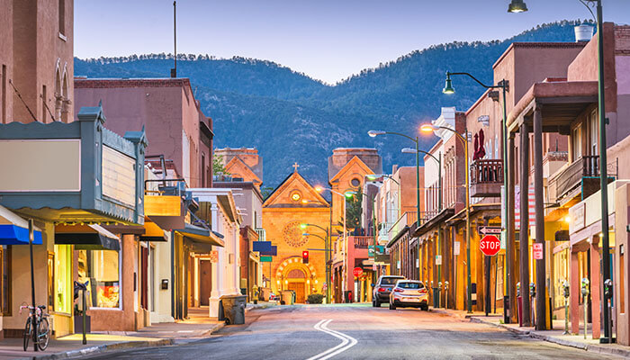 Charming evening view of Santa Fe, New Mexico, featuring adobe-style buildings, historic streets, and the illuminated Cathedral Basilica of St. Francis of Assisi with mountain scenery in the background.