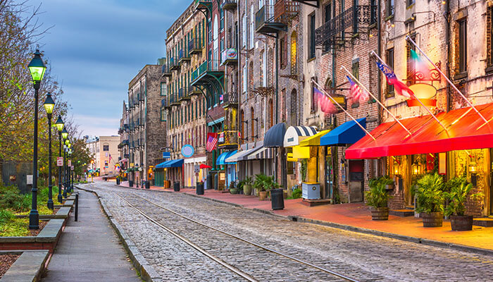 Historic River Street in Savannah, Georgia, lined with cobblestone streets, colorful shopfronts, and vintage brick buildings under soft evening light.