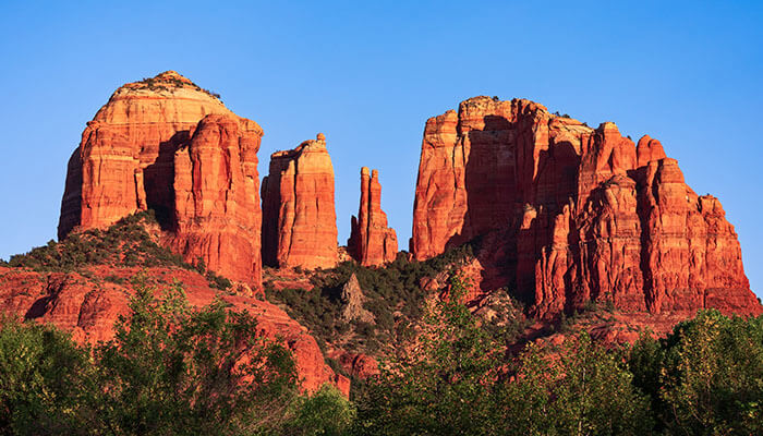 Majestic red rock formations of Cathedral Rock in Sedona, Arizona, glowing under clear blue skies with lush green vegetation at the base.
