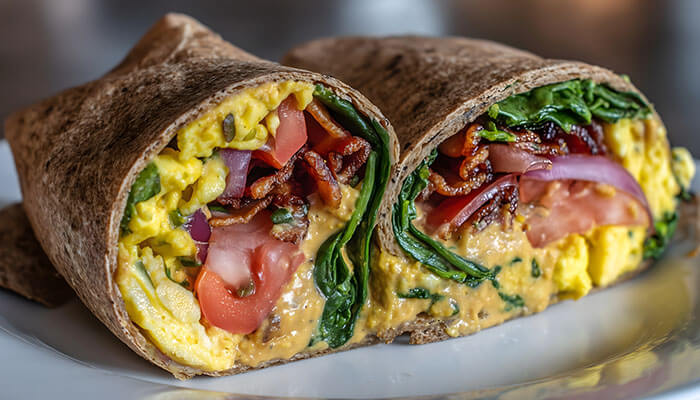 Close-up of a breakfast wrap cut in half, filled with scrambled eggs, tomatoes, spinach, bacon, and creamy sauce — a protein-packed morning meal.