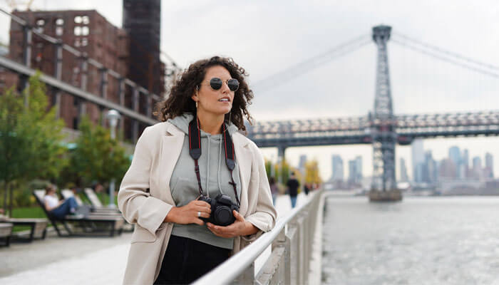 A solo female traveler holding a camera stands near a riverside walkway with a large bridge and city skyline in the background — one of the best places to travel solo female in US for photography and exploration.