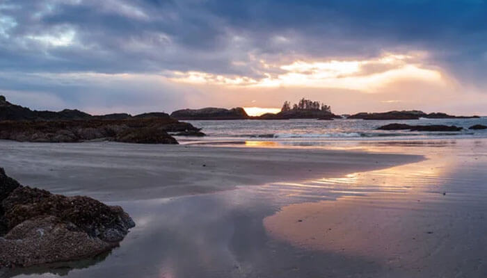 Sunset over the beach in Tofino, British Columbia, with reflections on wet sand and rocky islands in the distance.