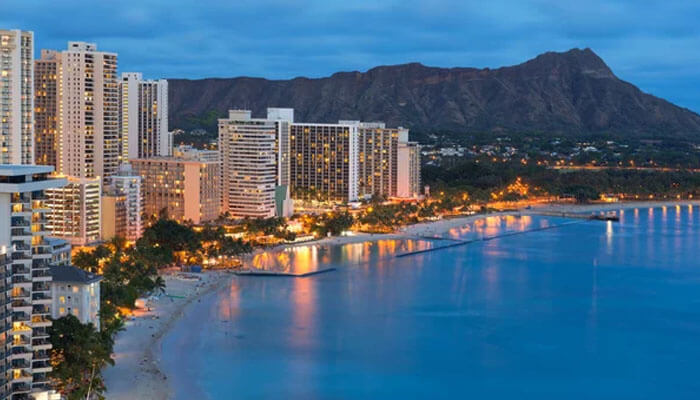Scenic view of Waikiki Beach in Honolulu, Hawaii, at dusk featuring illuminated beachfront hotels, calm ocean waters, and Diamond Head volcano in the distance.