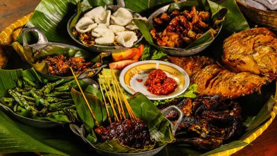 Traditional Indonesian food platter served on banana leaves featuring satay, fried chicken, sambal, grilled fish, and assorted side dishes.