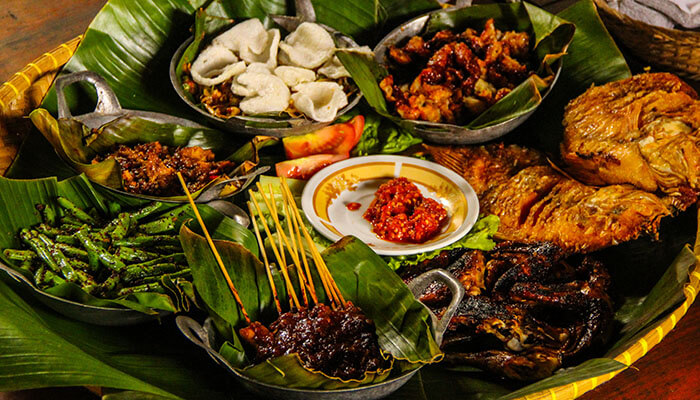 Traditional Indonesian food platter served on banana leaves featuring satay, fried chicken, sambal, grilled fish, and assorted side dishes.