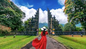 A person wearing a red dress and a wide-brimmed hat walking through the iconic gate at Lempuyang Temple, Bali, with lush greenery and a clear sky.