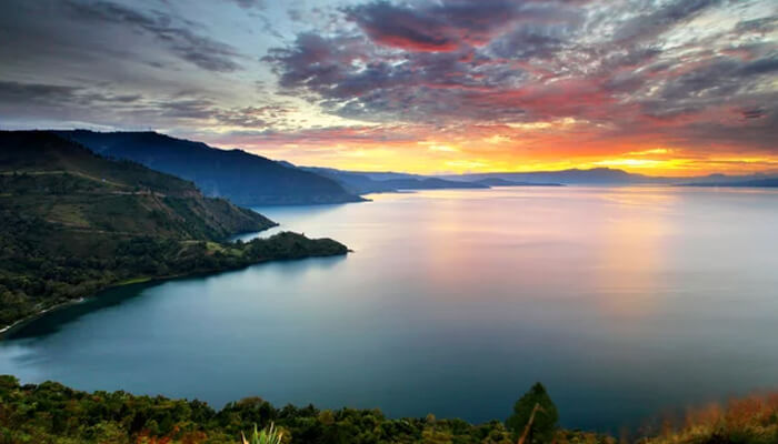 A stunning sunset over Lake Toba, Indonesia, with calm waters reflecting the colorful sky and lush greenery surrounding the lake.