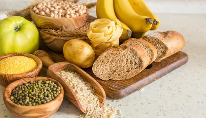 A selection of carbohydrate-rich foods including bananas, bread, pasta, rice, beans, potatoes, and grains on a wooden board
