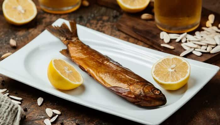 A smoked whole fish served on a white plate with lemon halves on the side