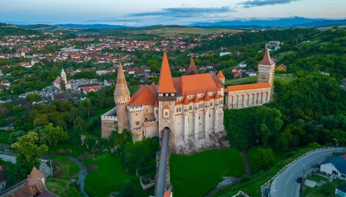 Best cities to visit in Romania-foodfuntrip - FoodFunTrip Aerial view of Corvin Castle in Romania, one of the cheapest European countries to visit, surrounded by lush green hills and traditional village houses, reflecting Romania's rich history and stunning landscapes.