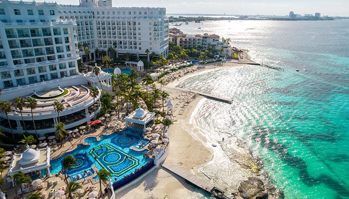 Riu Palace Peninsula - Food Fun Trip - FoodFunTrip An aerial view of a luxurious all-inclusive resort in Cancun, featuring a stunning beach, crystal-clear waters, and a large pool area surrounded by palm trees