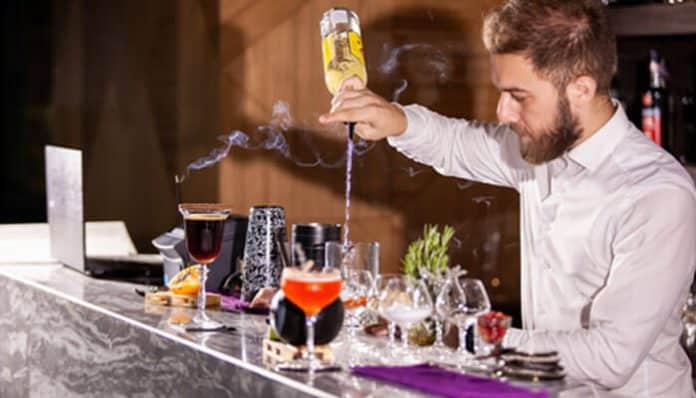 Bartender in a white shirt pouring liquor into a cocktail glass at a bar with multiple colorful drinks on the counter.