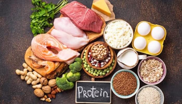 Building Around Lean Animal Protein - Food fun trip - FoodFunTrip Assorted high-protein foods on a wooden board, including meat, fish, eggs, dairy, legumes, nuts, and a chalk-sign reading 'Protein'.