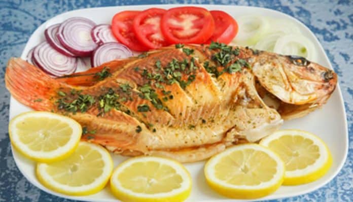 Grilled whole fish on a plate, garnished with herbs, with lemon slices, tomato, onion, and cucumber on the side.