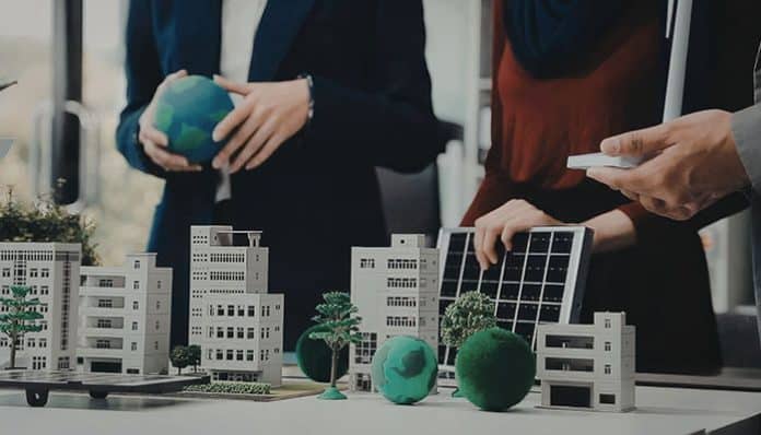 Team of professionals reviewing a sustainability-focused urban planning model with miniature buildings, trees, a globe, and a solar panel.