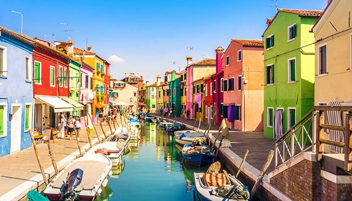 Visit the Colorful Island of Burano - Food Fun Trip - FoodFunTrip Colorful canal street with bright houses and small boats docked along the waterway (likely Burano). Possible view of a rainbow row of houses beside a calm canal with boats moored.