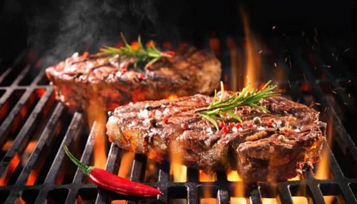 Two ribeye steaks cooking on a hot grill with rosemary sprigs and a red chili pepper nearby.