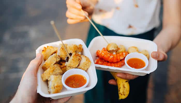 Ethical Food Travel and Sustainability - Food Fun Trip - FoodFunTrip Two hands hold white disposable trays with fried bite-sized pieces and dipping sauce; skewers are used to pick up the food, and a banana is visible under the second tray.
