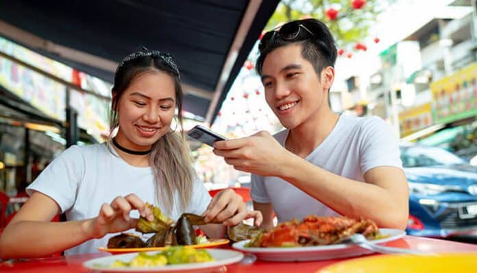 Two friends at an outdoor street market share plates of colorful dishes; one uses a smartphone to take a photo.
