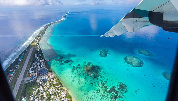 Is Yukevalo Island Safe - Food Fun Trip - FoodFunTrip Aerial view from an airplane window over a tropical coastline, with a turquoise reef and a small town along the shore and a visible wing in the top-right corner.