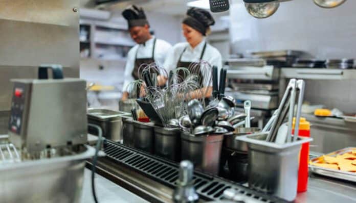 Two chefs in white uniforms and black hats work in a busy commercial kitchen, sorting metal utensils by the sink area.