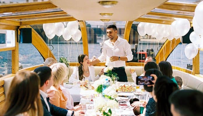 Man giving a toast at a wedding reception on a boat, guests seated along a long table with white balloons and flowers nearby.