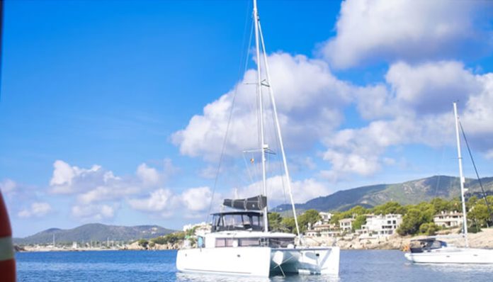 White catamaran anchored in calm blue water near a coastal town, with green hills and white buildings in the distance under a sunny sky.
