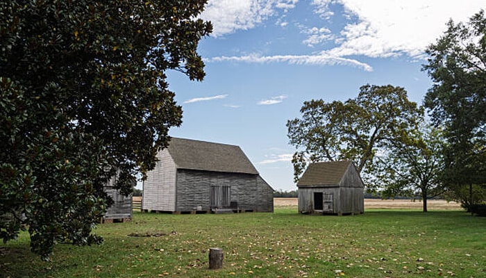 John Dickinson Plantation - Food Fun Trip - FoodFunTrip Three weathered wooden farm buildings sit on a green field under a bright blue sky with scattered clouds and trees nearby.