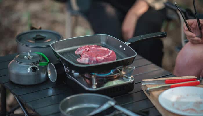 Kitchen Necessities - Food Fun Trip - FoodFunTrip Close-up of a Caravan Adventure campsite cooking setup featuring a portable gas stove with a frying pan searing fresh steak, surrounded by camping cookware such as a kettle, pots, and utensils, highlighting practical outdoor meal preparation during off-grid travel.