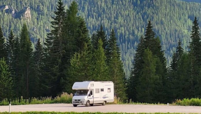 A white camper van parked on a roadside with tall evergreen trees and forested hills in the background.