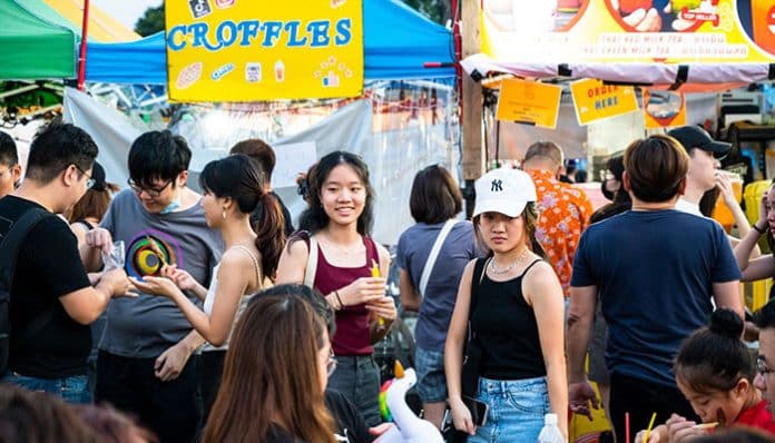 Crowded outdoor festival scene with colorful tents, banners, and people chatting and enjoying food and drinks at a market stall row.