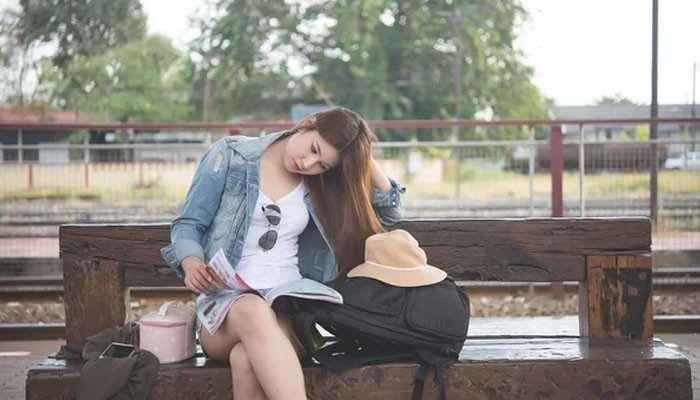 Take Solo Travel in Manageable Steps - Food Fun Trip - FoodFunTrip Backpacking Tips CWBiancaVoyage From ConversationsWithBianca featuring a solo female traveler reading a guidebook at a train station with her backpack and travel hat.